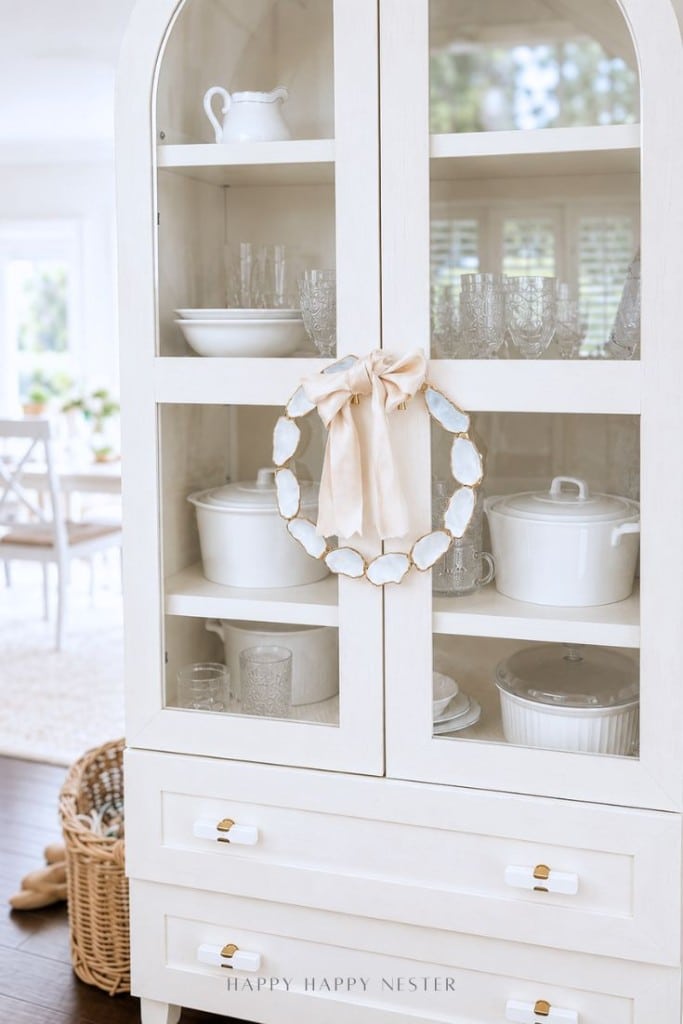 A white china cabinet with glass doors displays white dishware and glassware. An oyster shell wreath DIY with a beige ribbon hangs on the door. The room is bright with natural light, and a basket sits on the floor beside the cabinet.