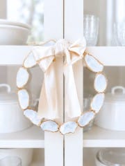 A decorative oyster shell wreath DIY, crafted from oval white stones with gold edges, is adorned with a large pale satin bow and hangs on a white cabinet door. White dishes and glassware are displayed inside the cabinet.