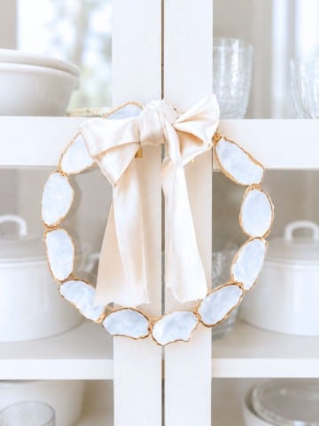A decorative oyster shell wreath DIY, crafted from oval white stones with gold edges, is adorned with a large pale satin bow and hangs on a white cabinet door. White dishes and glassware are displayed inside the cabinet.
