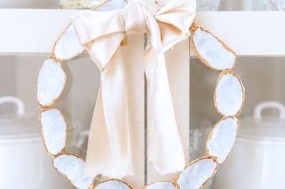 A decorative oyster shell wreath DIY, crafted from oval white stones with gold edges, is adorned with a large pale satin bow and hangs on a white cabinet door. White dishes and glassware are displayed inside the cabinet.
