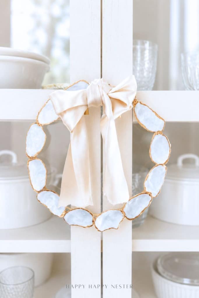 A decorative oyster shell wreath DIY, crafted from oval white stones with gold edges, is adorned with a large pale satin bow and hangs on a white cabinet door. White dishes and glassware are displayed inside the cabinet.