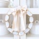 A decorative oyster shell wreath DIY, crafted from oval white stones with gold edges, is adorned with a large pale satin bow and hangs on a white cabinet door. White dishes and glassware are displayed inside the cabinet.