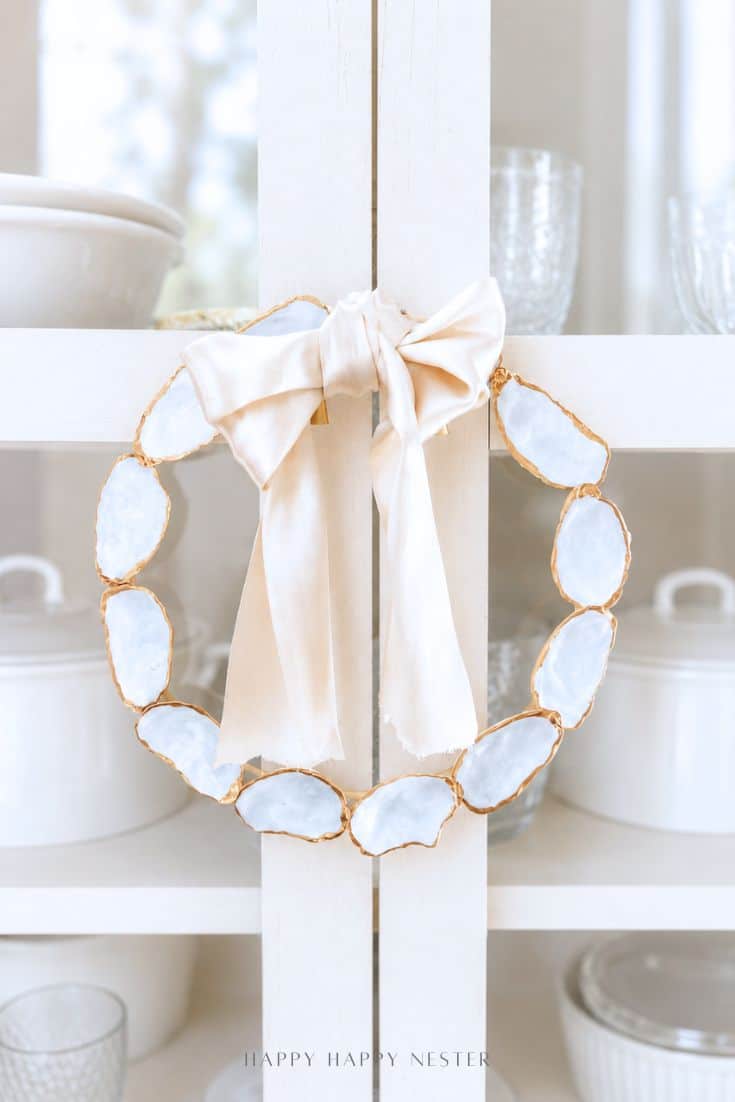 A decorative oyster shell wreath DIY, crafted from oval white stones with gold edges, is adorned with a large pale satin bow and hangs on a white cabinet door. White dishes and glassware are displayed inside the cabinet.