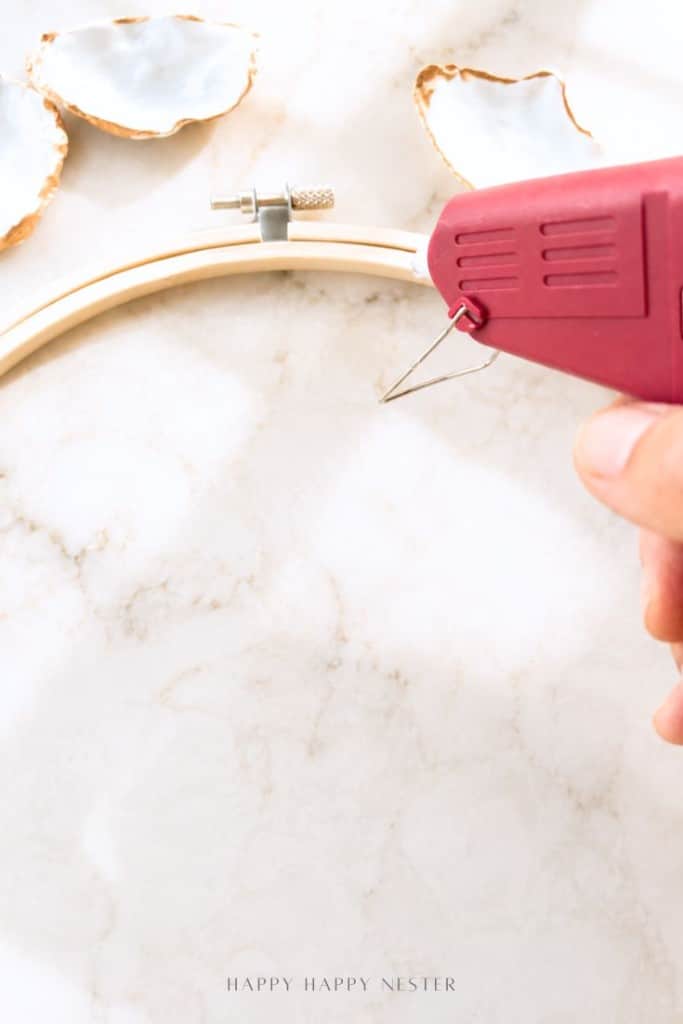 A hand holds a red hot glue gun, applying glue to a wooden embroidery hoop on a marble surface. Decorative oyster shells with gold edges are nearby.