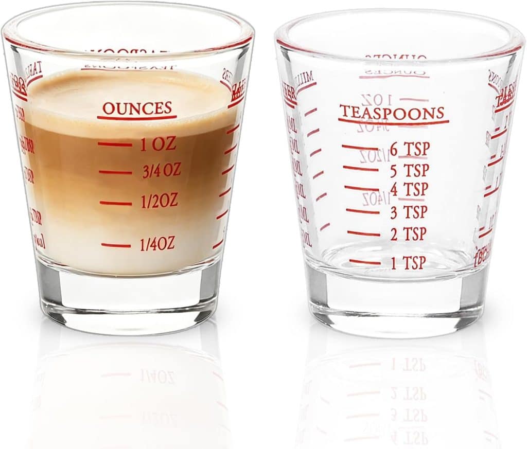 Two clear glass measuring cups—one filled with a layered espresso drink up to the 1-ounce mark, the other empty—feature red measurement markings for ounces, tablespoons, and teaspoons. Perfect for crafting precise drinks or testing out a ginger beer mocktail recipe.