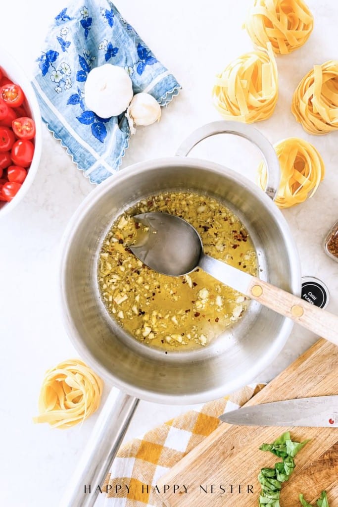 Overhead view of a pot on a counter with chopped garlic cooking in oil. Surrounding the pot are uncooked pasta nests, tomatoes, a blue cloth, garlic cloves, and a wooden cutting board with chopped herbs.