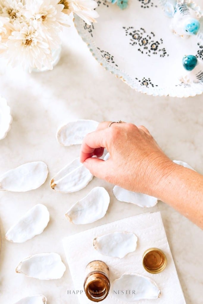 A hand paints the edge of white oyster shells with gold paint on a napkin, surrounded by shells, a small paint jar, white flowers, and a decorative plate on a marble surface.