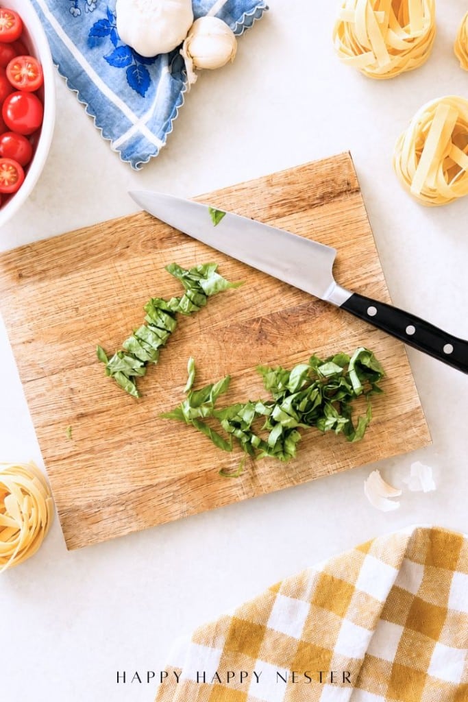 A wooden cutting board with chopped basil and a knife on top, surrounded by raw pasta nests, a bowl of cherry tomatoes, mushrooms, and a blue and yellow checkered cloth—perfect ingredients for a simple pasta sauce recipe.