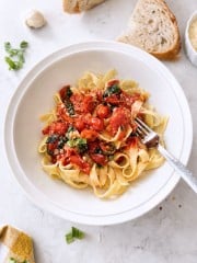 Simple Pasta Sauce Recipe A bowl of tagliatelle pasta topped with cherry tomatoes, spinach, and grated cheese highlights a simple pasta sauce recipe. A fork rests in the bowl, while slices of bread, garlic, and fresh basil are arranged on a marble surface nearby.