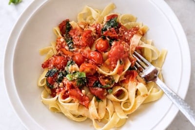 A bowl of tagliatelle pasta topped with cherry tomatoes, spinach, and grated cheese highlights a simple pasta sauce recipe. A fork rests in the bowl, while slices of bread, garlic, and fresh basil are arranged on a marble surface nearby.