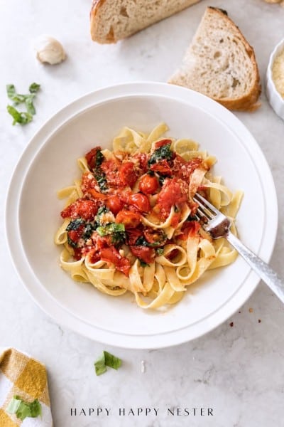 A bowl of tagliatelle pasta topped with cherry tomatoes, spinach, and grated cheese highlights a simple pasta sauce recipe. A fork rests in the bowl, while slices of bread, garlic, and fresh basil are arranged on a marble surface nearby.