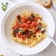 A bowl of tagliatelle pasta topped with cherry tomatoes, spinach, and grated cheese highlights a simple pasta sauce recipe. A fork rests in the bowl, while slices of bread, garlic, and fresh basil are arranged on a marble surface nearby.