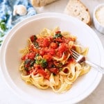 A white bowl of tagliatelle pasta topped with a simple pasta sauce recipe and basil sits with a fork inside. Surrounding the bowl are slices of bread, parmesan, a garlic clove, a blue napkin, and fresh basil leaves.