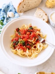 A white bowl of tagliatelle pasta topped with a simple pasta sauce recipe and basil sits with a fork inside. Surrounding the bowl are slices of bread, parmesan, a garlic clove, a blue napkin, and fresh basil leaves.