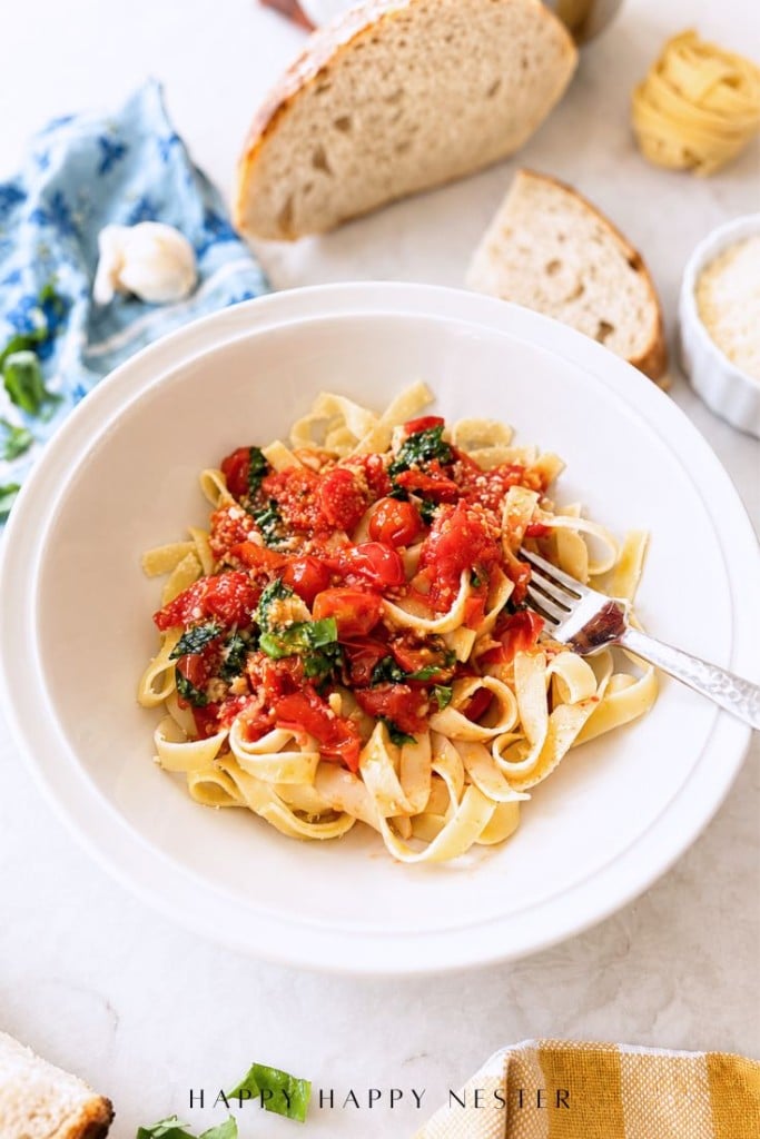 A white bowl of tagliatelle pasta topped with a simple pasta sauce recipe and basil sits with a fork inside. Surrounding the bowl are slices of bread, parmesan, a garlic clove, a blue napkin, and fresh basil leaves.