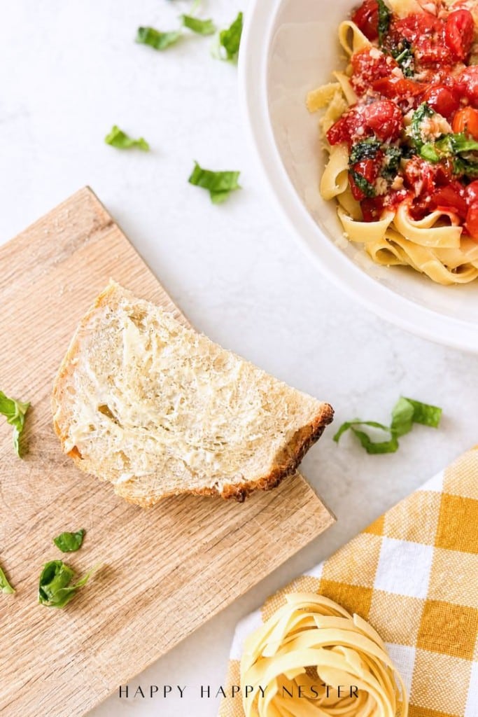 A slice of buttered bread on a wooden board sits next to a bowl of pasta with tomato sauce and herbs, featuring fresh basil leaves and a yellow checkered napkin—the perfect setting for enjoying a simple pasta sauce recipe.