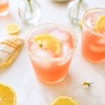 A pink ginger beer mocktail recipe with ice and a lemon slice in a sugar-rimmed glass, surrounded by lemon wedges, a striped napkin, and vases of pink and yellow flowers on a light surface.