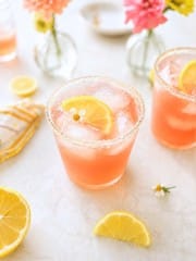 A pink ginger beer mocktail recipe with ice and a lemon slice in a sugar-rimmed glass, surrounded by lemon wedges, a striped napkin, and vases of pink and yellow flowers on a light surface.