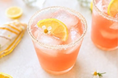 A pink ginger beer mocktail recipe with ice and a lemon slice in a sugar-rimmed glass, surrounded by lemon wedges, a striped napkin, and vases of pink and yellow flowers on a light surface.