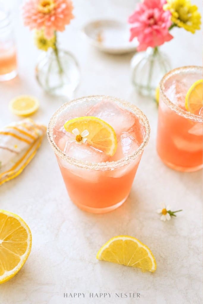 A pink ginger beer mocktail recipe with ice and a lemon slice in a sugar-rimmed glass, surrounded by lemon wedges, a striped napkin, and vases of pink and yellow flowers on a light surface.