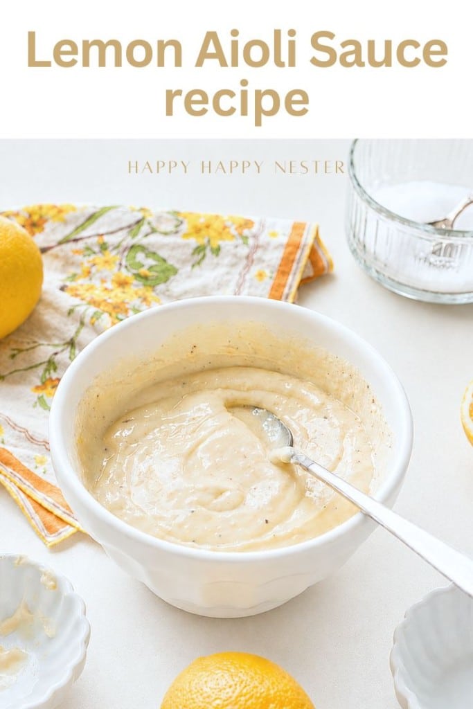 A bowl of creamy lemon aioli sits with a spoon, surrounded by lemons, a floral napkin, and small bowls. The text "Lemon Aioli Sauce Recipe" above highlights this easy lemon aioli sauce recipe perfect for any dish.