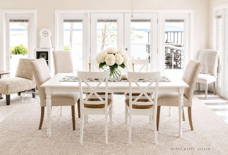 Bright dining room with a white table, beige upholstered chairs, two white chairs with woven seats, and a centerpiece of white flowers. Large windows in the background overlook water and greenery.
