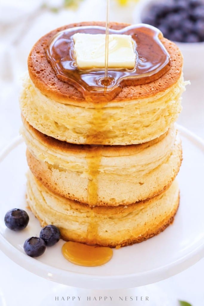 A stack of thick, fluffy pancakes topped with a pat of butter and syrup being poured over them, with a few blueberries and syrup on the plate.