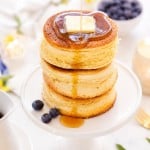 A stack of three fluffy pancakes topped with a pat of butter is being drizzled with syrup. The pancakes sit on a white pedestal plate, surrounded by blueberries and a bright, inviting breakfast setting.