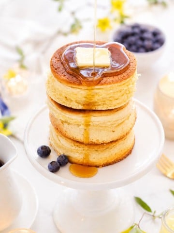 A stack of three fluffy pancakes topped with a pat of butter is being drizzled with syrup. The pancakes sit on a white pedestal plate, surrounded by blueberries and a bright, inviting breakfast setting.