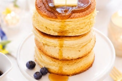 A stack of three fluffy pancakes topped with a pat of butter is being drizzled with syrup. The pancakes sit on a white pedestal plate, surrounded by blueberries and a bright, inviting breakfast setting.