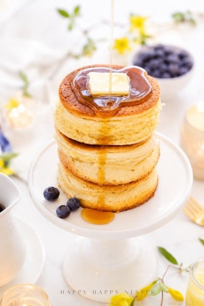 A stack of three fluffy pancakes topped with a pat of butter is being drizzled with syrup. The pancakes sit on a white pedestal plate, surrounded by blueberries and a bright, inviting breakfast setting.