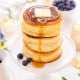 A stack of three fluffy pancakes topped with a pat of butter is being drizzled with syrup. The pancakes sit on a white pedestal plate, surrounded by blueberries and a bright, inviting breakfast setting.