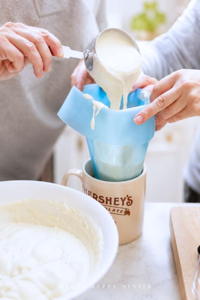 Two people pour cake batter through a blue silicone funnel into a Hershey’s mug, with a bowl of batter nearby on a kitchen counter.