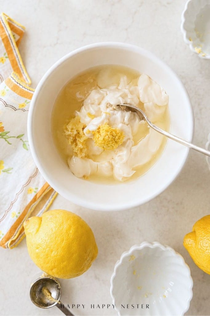 A white bowl containing mayonnaise, lemon juice, and lemon zest being mixed with a spoon. Lemons, a measuring spoon, and decorative napkin are nearby on a light-colored countertop.