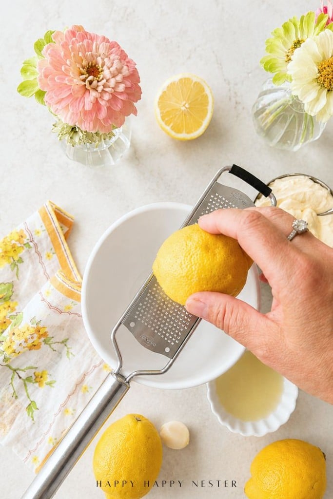 A hand zests a lemon over a white bowl with a metal grater—perfect prep for a fresh lemon aioli sauce recipe. Lemons, lemon juice, flowers in vases, and a floral napkin are arranged on a light surface nearby.