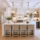 Bright, modern kitchen with white cabinetry, a large island with four woven stools, gold pendant lights, stainless steel appliances, and large windows letting in natural light, with greenery and decorative accents throughout.