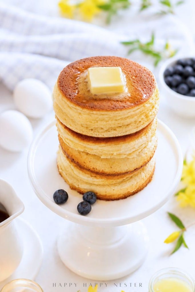 Homemade Japanese soufflé pancake recipe served for breakfast pancakes topped with a pat of butter, served on a white cake stand with blueberries on the side. Eggs, blueberries, and flowers are blurred in the background.