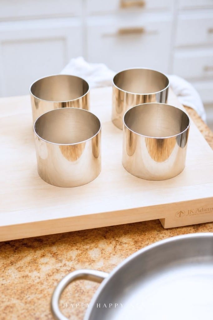 Four stainless steel ring molds are arranged on a wooden cutting board in a kitchen setting, with a metal pan partially visible in the foreground. White cabinets and drawers are in the background.
