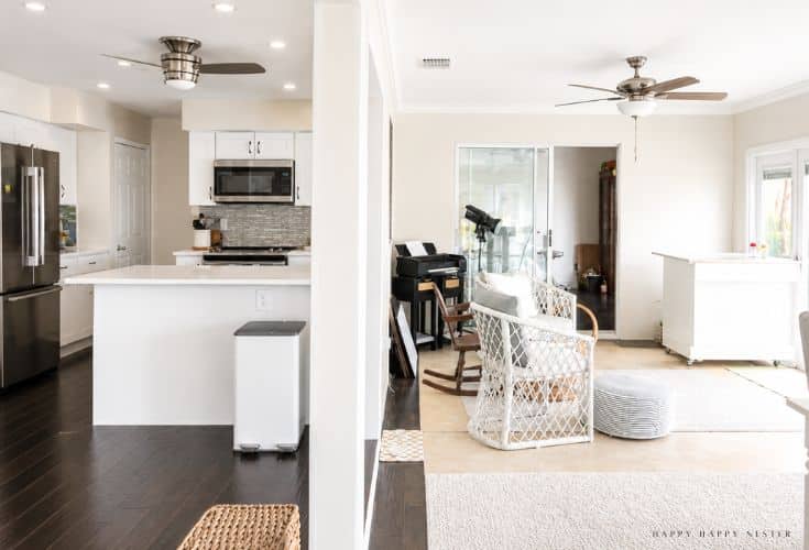Bright open-concept home featuring a modern white kitchen with stainless steel appliances and a connected living area with wicker chairs, a keyboard, and large windows letting in natural light.