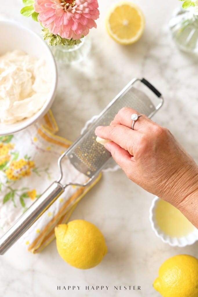 A hand with a ring grates lemon zest over a metal grater on a marble surface, surrounded by whole and halved lemons, a bowl of cream, a lemon-patterned cloth, and a pink flower.