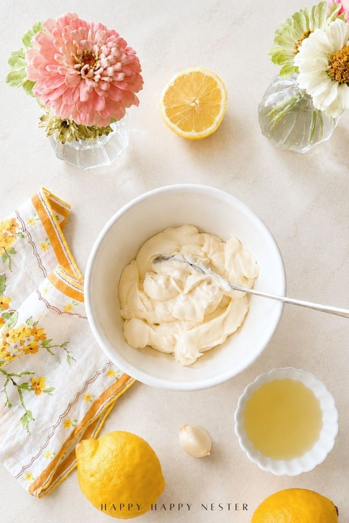 A bowl of creamy lemon aioli sauce, with a spoon, surrounded by lemons, lemon juice, a garlic clove, a floral napkin, and two small vases of flowers on a light surface—the perfect setup for your next lemon aioli sauce recipe.