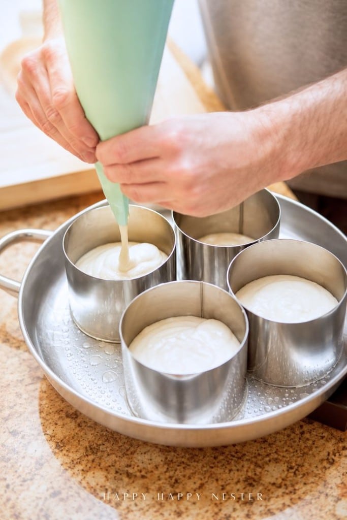 A person pipes batter into round metal baking rings placed inside a large pan on a kitchen counter, preparing individual cakes for baking.