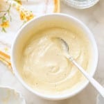 A white bowl filled with creamy, light yellow lemon aioli sauce rests on a countertop, a spoon inside. Nearby are a floral napkin, a small bowl, and a glass bowl of salt—perfect for your next lemon aioli sauce recipe.