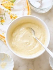 A white bowl filled with creamy, light yellow lemon aioli sauce rests on a countertop, a spoon inside. Nearby are a floral napkin, a small bowl, and a glass bowl of salt—perfect for your next lemon aioli sauce recipe.