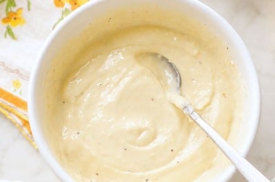 A white bowl filled with creamy, light yellow lemon aioli sauce rests on a countertop, a spoon inside. Nearby are a floral napkin, a small bowl, and a glass bowl of salt—perfect for your next lemon aioli sauce recipe.
