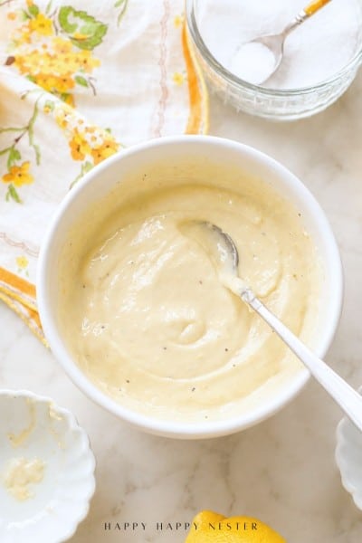 A white bowl filled with creamy, light yellow lemon aioli sauce rests on a countertop, a spoon inside. Nearby are a floral napkin, a small bowl, and a glass bowl of salt—perfect for your next lemon aioli sauce recipe.