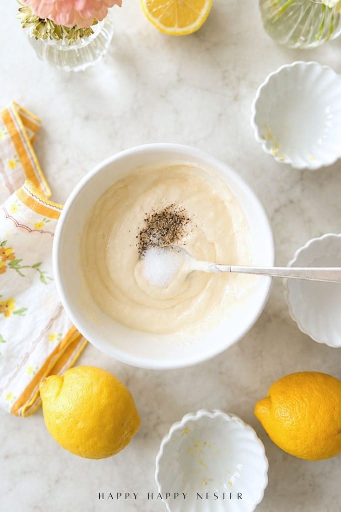 A bowl of creamy lemon aioli sauce recipe with salt and pepper on top, surrounded by lemons, empty scalloped bowls, a floral napkin, and a vase with flowers on a light countertop.