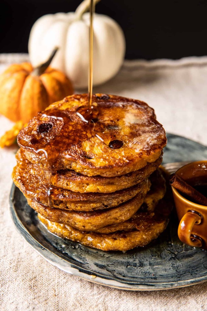 A stack of pumpkin pancakes on a plate is drizzled with syrup, with small pumpkins and a cup of syrup in the background.