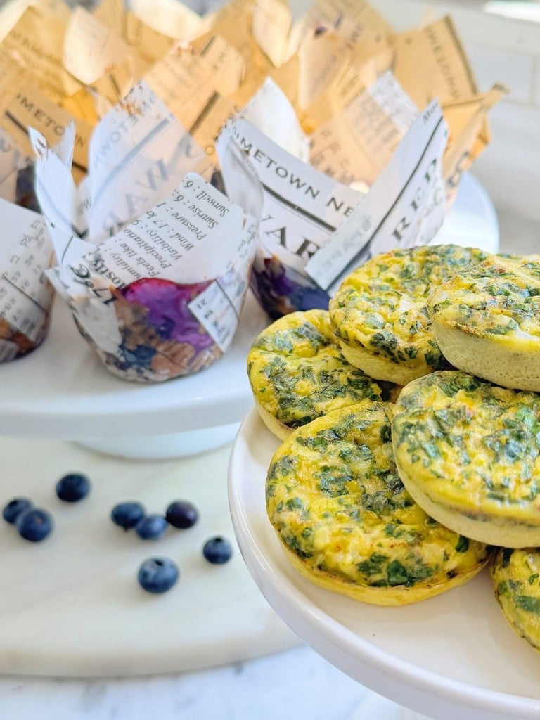 A plate of mini spinach and egg frittatas sits in front, with blueberry muffins wrapped in newspaper-style paper in the background. Fresh blueberries are scattered on a white surface nearby.