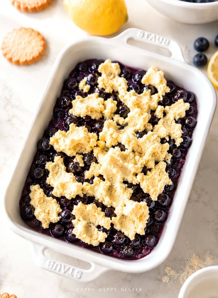 A white baking dish filled with blueberries, topped with dollops of crumbly biscuit dough, sits on a light countertop with a lemon, cookies, and blueberries nearby.